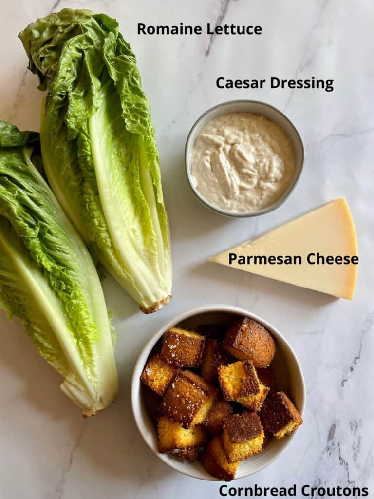 Ingredients needed for the Caesar salad. Pictured ingredients needed are romaine lettuce, cornbread croutons, parmesan cheese and creamy Caesar dressing.