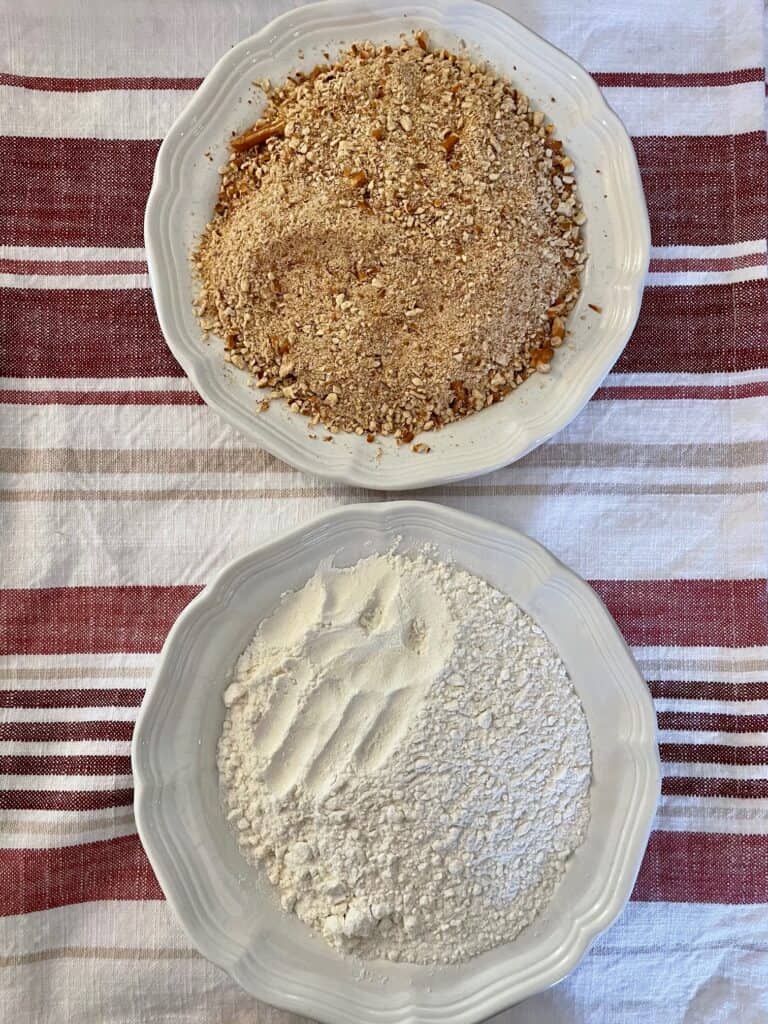 Photo is showing two plates. The first plate contains the pretzel crumbs and the second plate contains the all purpose flour. 