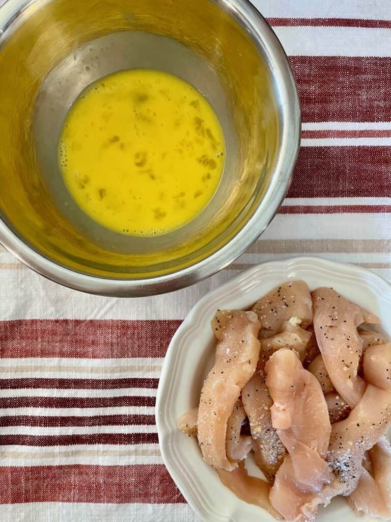 Photo contains a small bowl with 2 eggs beaten in the bowl and a plate with the raw chicken tenders. The chicken tenders are sprinkled with salt, pepper and garlic powder. 