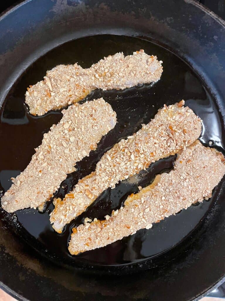 This photo shows the pretzel crusted chicken tenders frying in a cast iron pan before they have been flipped over.
