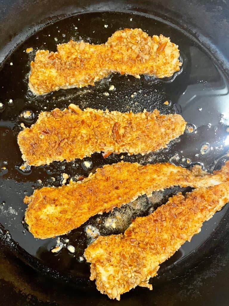 Photo shows the pretzel crusted chicken tenders frying in a cast iron pan after they have been flipped once.