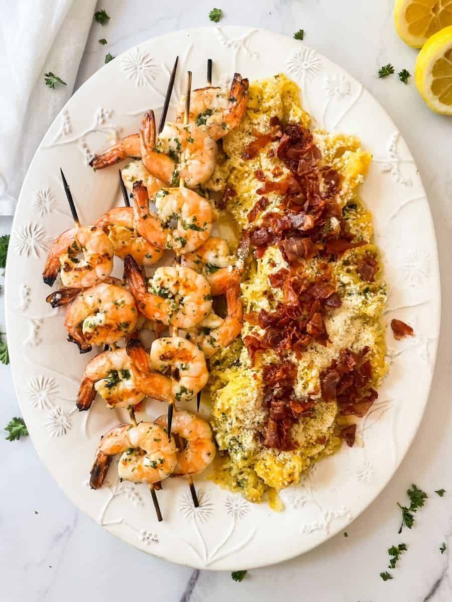 Shrimp and spaghetti squash displayed on a platter. Spaghetti squash is topped with parmesan cheese and crispy prosciutto. Shrimp is on skewers.