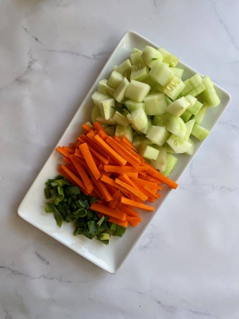 Raw veggies for the sushi bowl chopped and displayed on a rectangular white plate. The image shows chopped cucumbers, carrots cut into match sticks and diced green onion. 