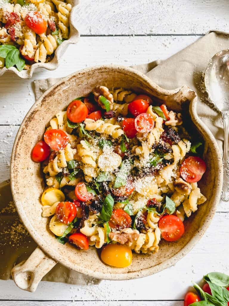 Pesto Pasta Salad with Sundried Tomatoes served in a tan colored pottery bowl. A small portion in a cream colored pottery bowl is featured in the top left corner. The bottom right corner shows a few cherry tomatoes and fresh basil leaves.