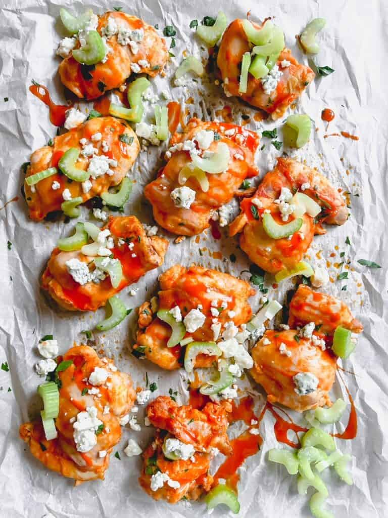 Buffalo chicken thigs on a sheet pan and parchment paper topped with crumbly blue cheese and chopped celery.