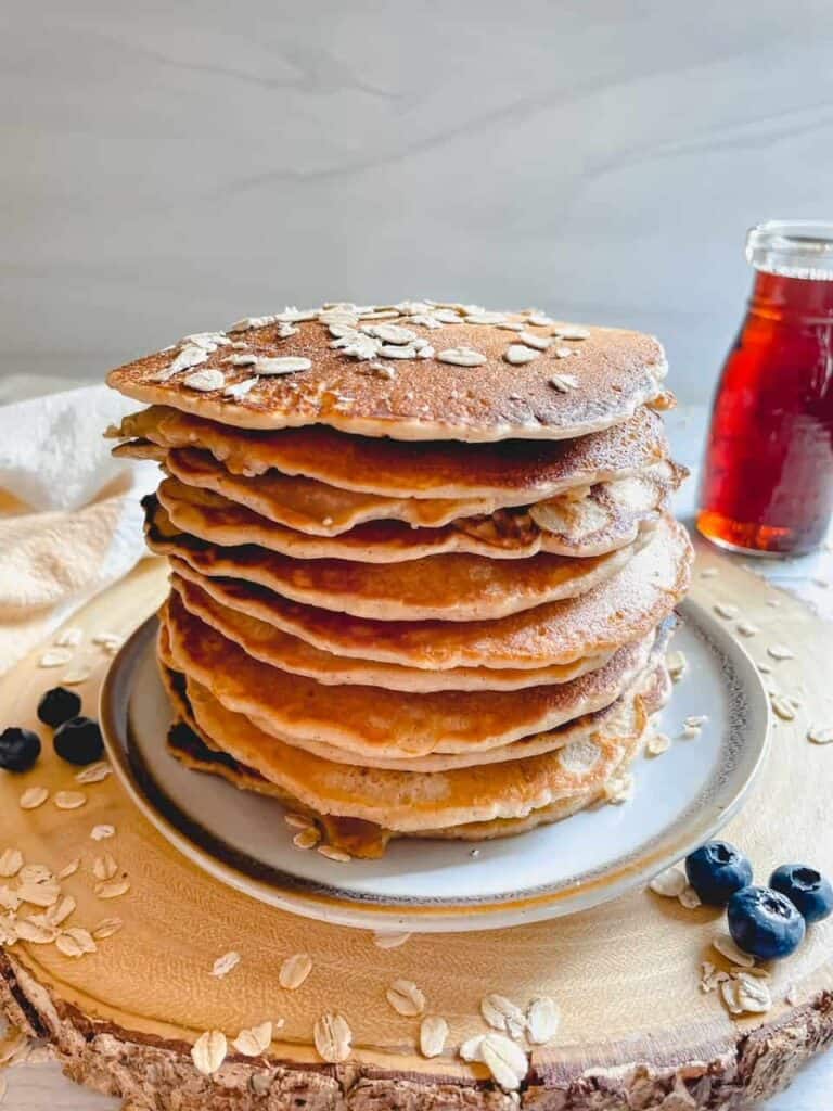 Stack of oat flour pancakes on a round wooden board with dry oats sprinkled around and a few fresh blueberries.