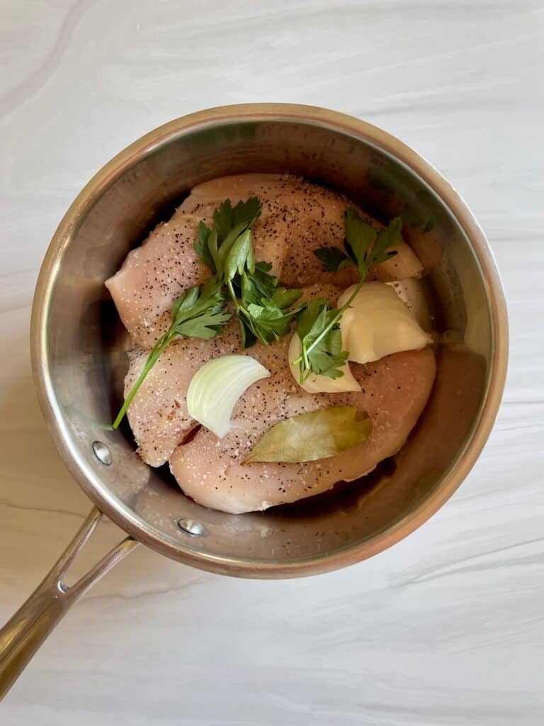 Raw chicken breast topped with white onion, a bay leaf and fresh parsley are in a saucepan.