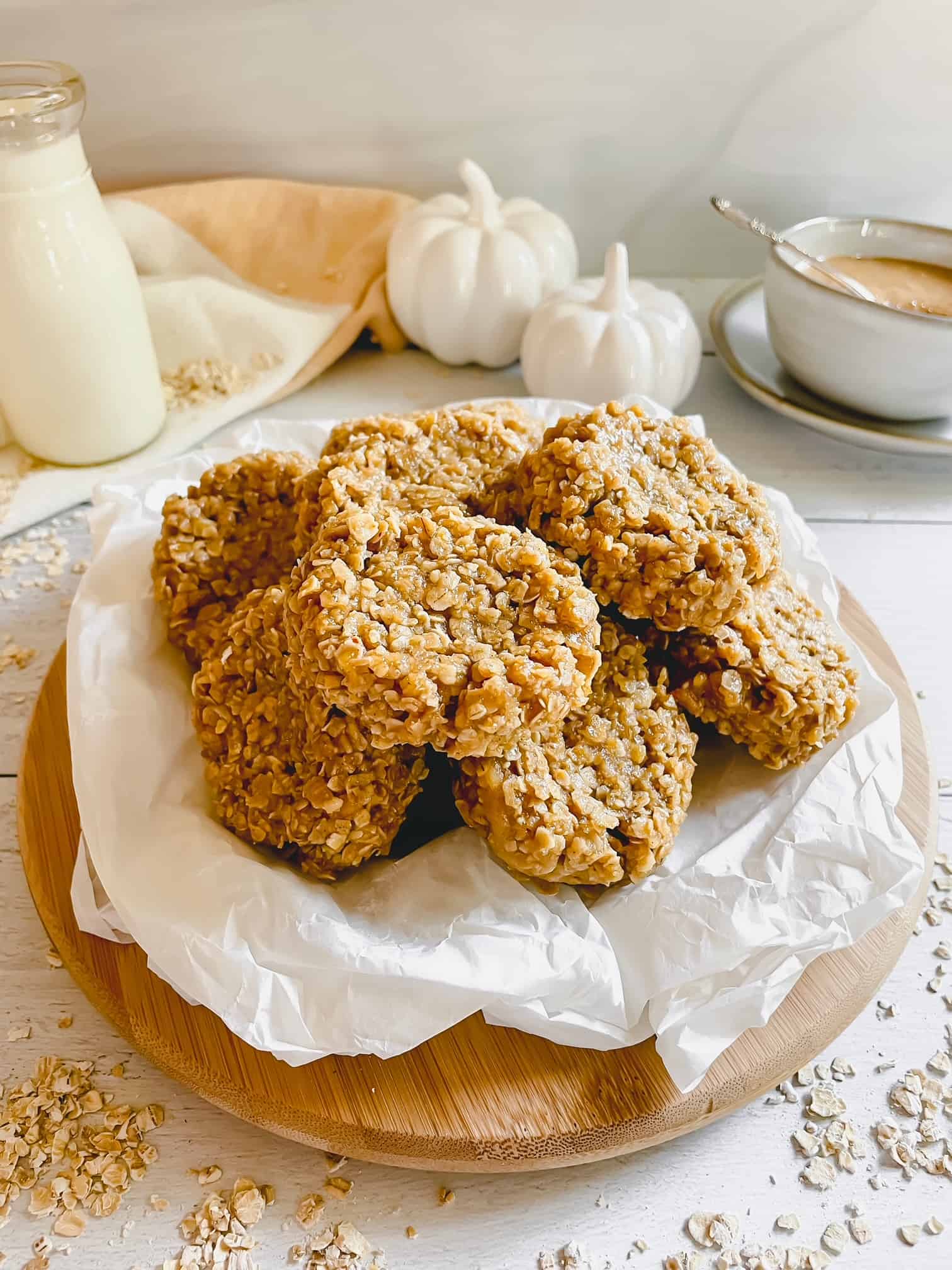 No bake cookies displayed on parchment paper and a round wooden cutting board. A glass of milk is in the background. Two white ceramic pumpkins are also in the background. A small bowl of peanut butter with a spoon can also be seen in the back of the image.