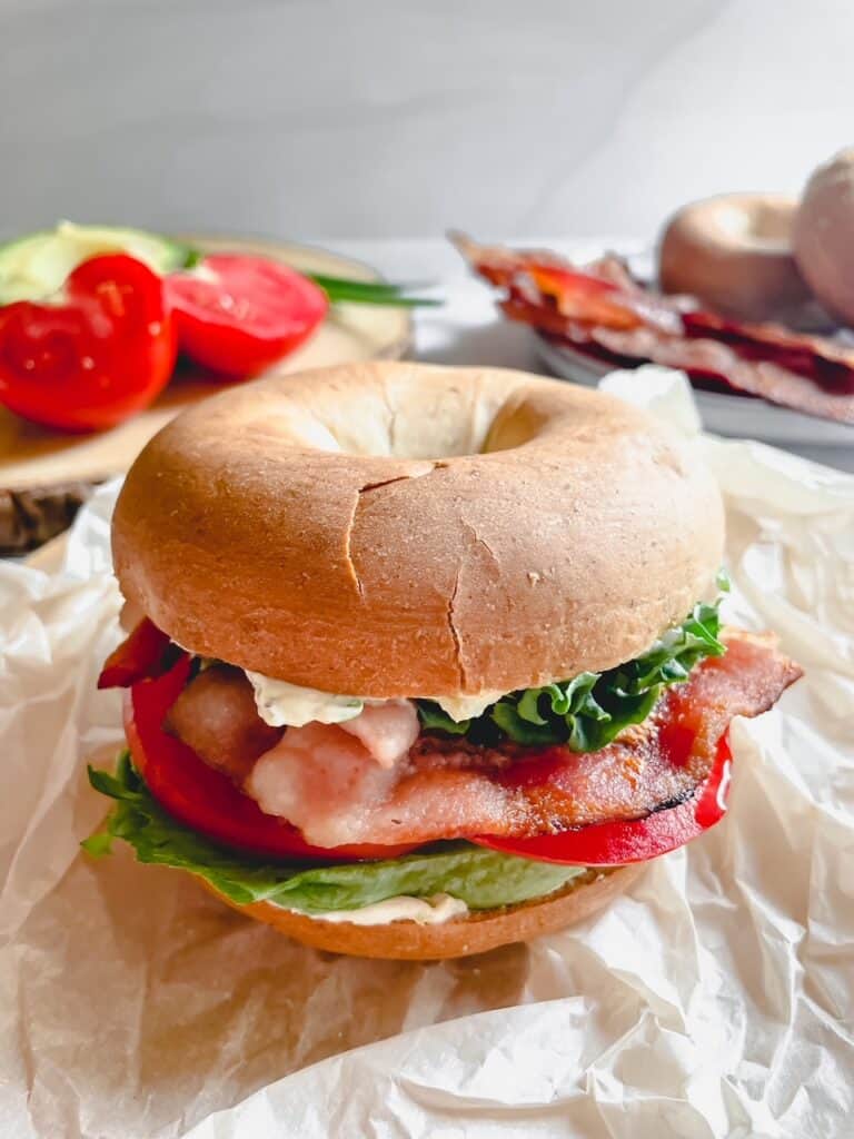 BLT Bagel on parchment paper with extra ingredients in the background.