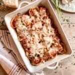 Cooked meatball stuffed shells in a white casserole dish with handles. A chunk of parmesan cheese, an antique cheese grater, a small wooden bowl with marinara sauce, fresh parsley, a head of garlic and a kitchen towel surround the dish of pasta.