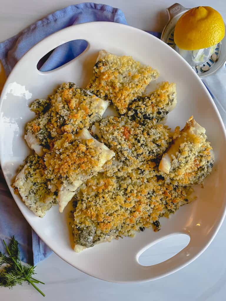 Panko cod filets on a white oval serving dish sat on top of a blue napkin. A lemon is in the top right corner. Fresh dill is in the bottom left corner.