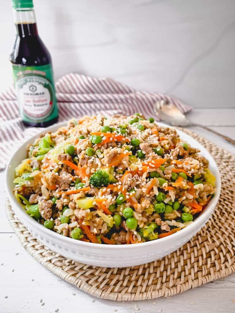 Teriyaki turkey, vegetables and rice in a large, shallow white bowl. A bottle of soy sauce is in the background on a red and white striped napkin.