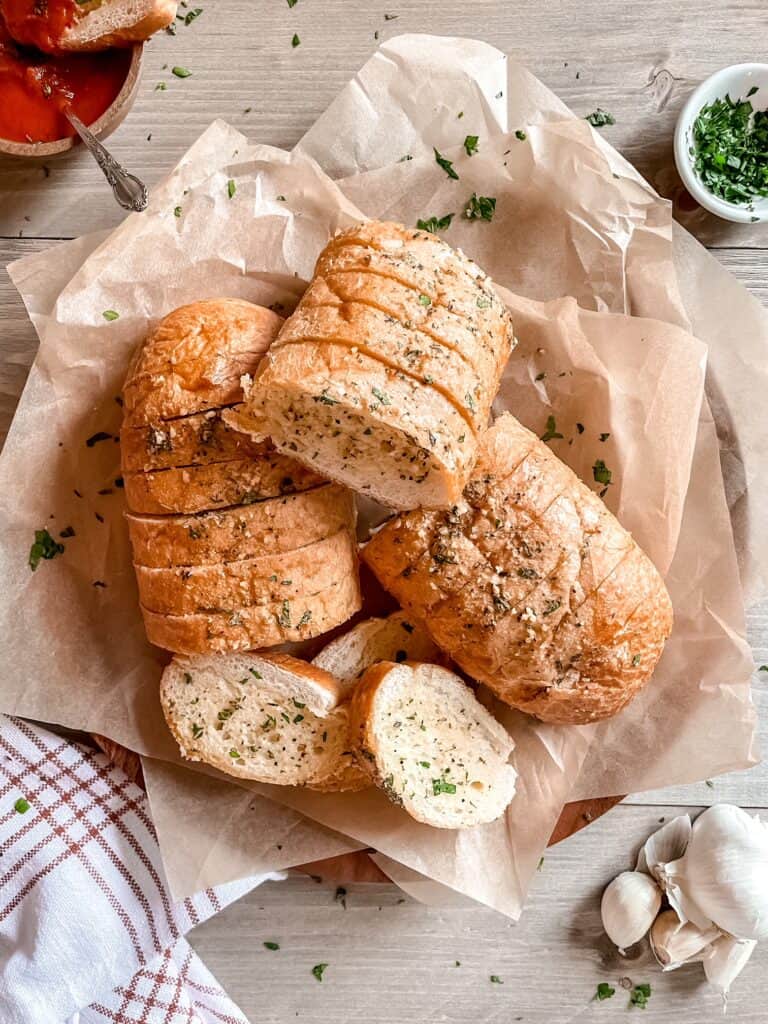 Vegan garlic bread on parchment paper on top of a brown, round cutting board. Marinara sauce in a wooden bowl in the top left corner. Fresh garlic is in the bottom right corner. Fresh diced parsley is in the top right corner in a small white bowl. A white and red striped kitchen towel is in the bottom left corner.