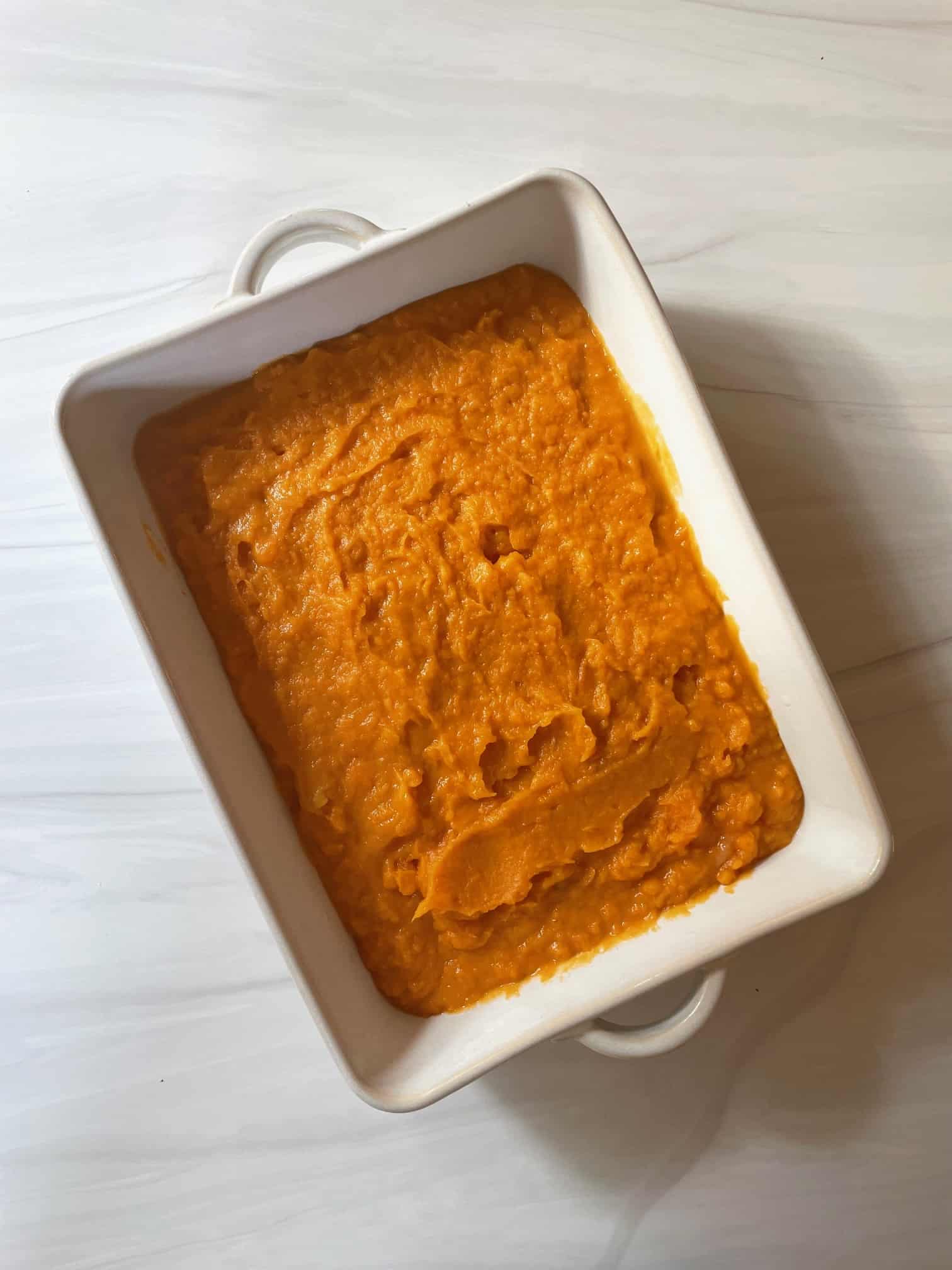 Mashed sweet potatoes mixed with liquid ingredients spread out in a white casserole dish