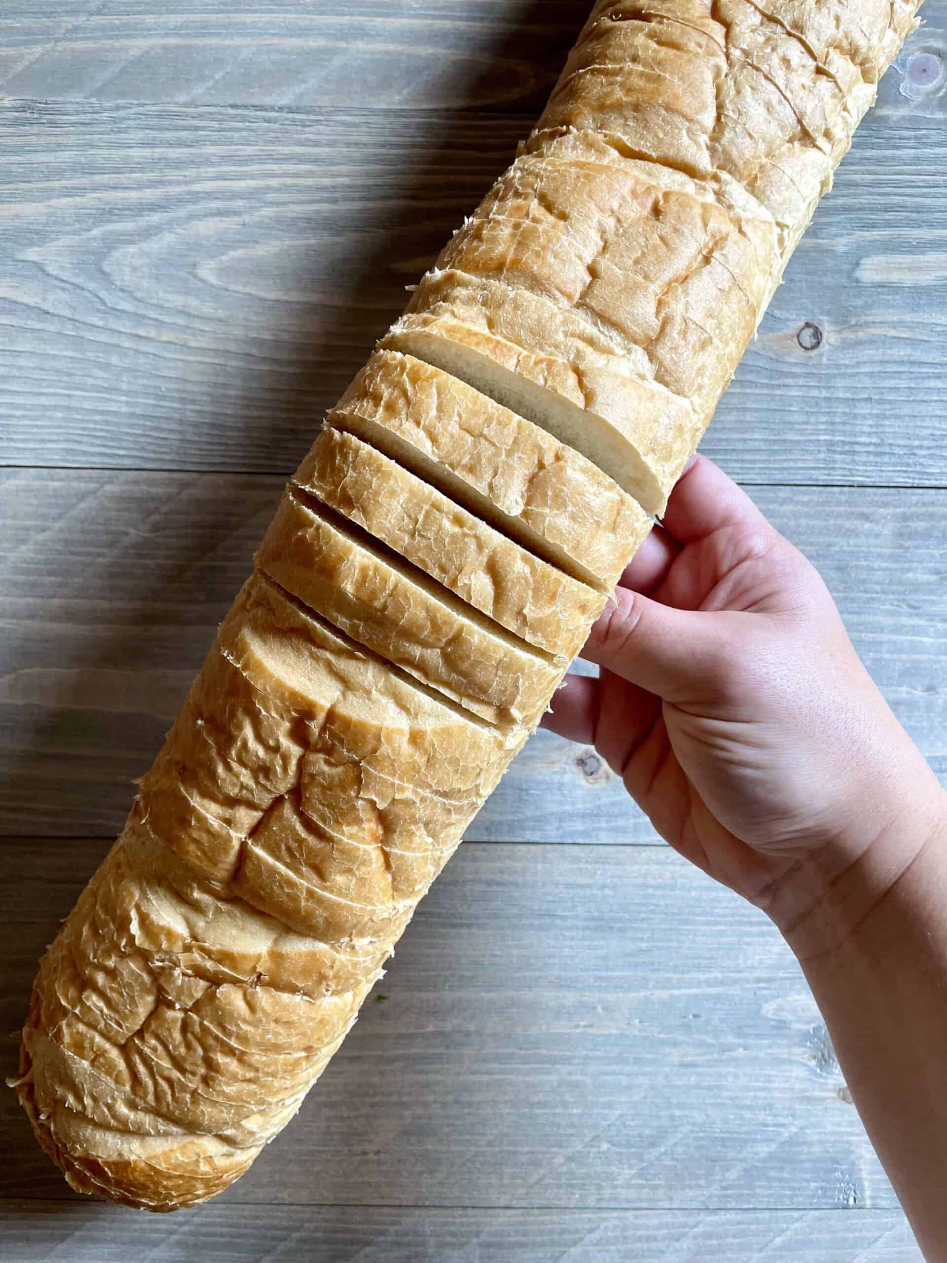 French bread is shown with slices cut into it. The bread is held up by a hand.