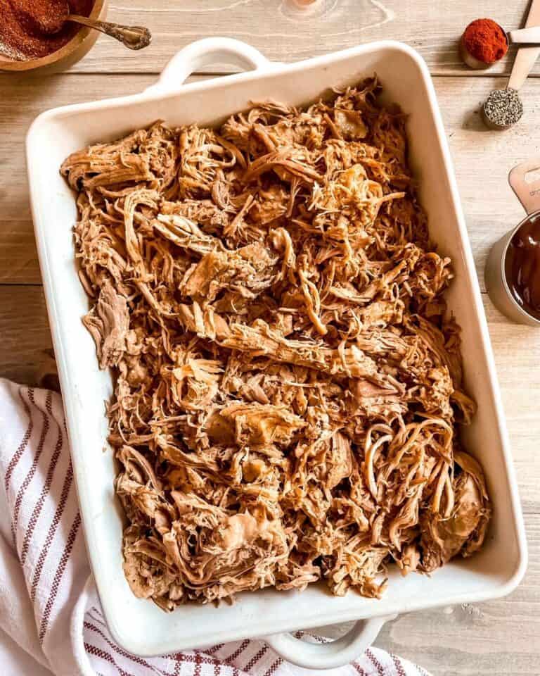 Pulled pork is in a white casserole dish, a red and white striped dish cloth is in the bottom left corner, spices are in the top corners, bbq sauce is on the side