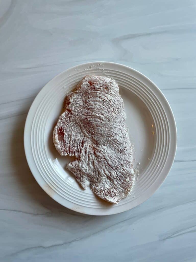 Chicken cutlet on a white plate coated in flour.