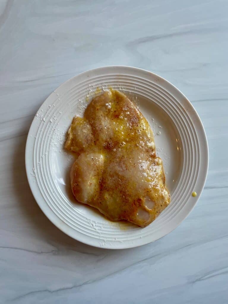 chicken breast piece on a white plate coated in an egg wash.