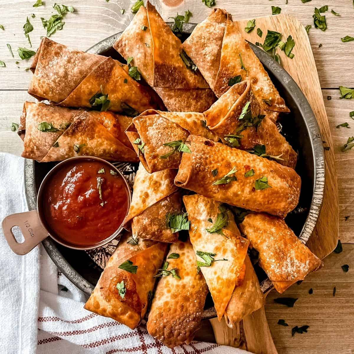 air fryer pizza rolls cooked, in a silver round pie plate, with marinara sauce on the side, garnished with parsley, red and white striped towel in the bottom left corner
