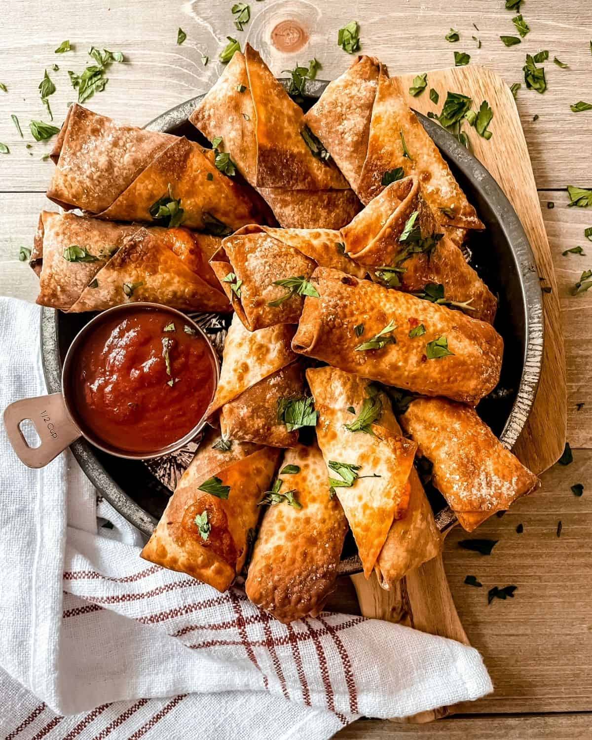 air fryer pizza rolls in a silver round pie plate, on a wooden cutting board, garnished with parsley, with a red and white striped dish towel in the bottom left corner