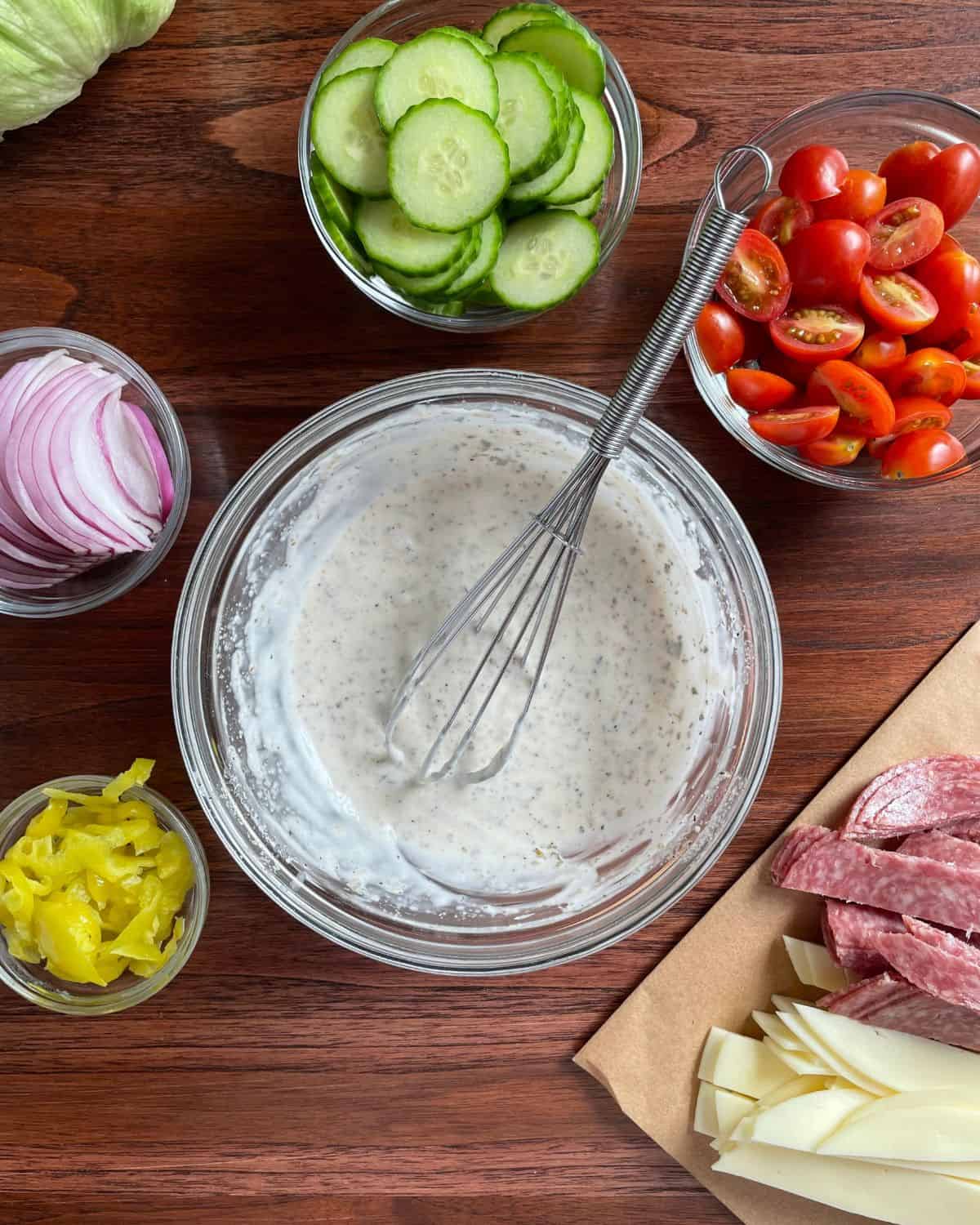 Creamy italian dressing in a glass bowl with a whisk, surrounded by various salad ingredients.