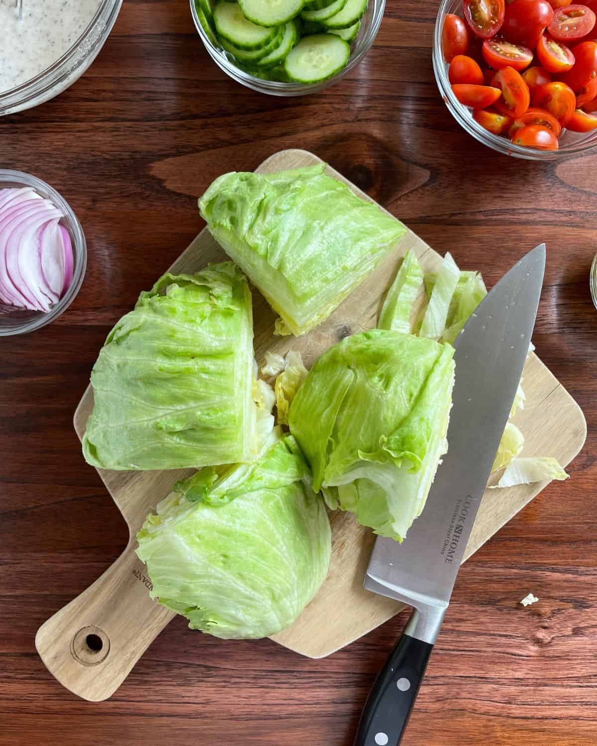 Iceberg lettuce chopped into wedges, with a knife and various vegetables.
