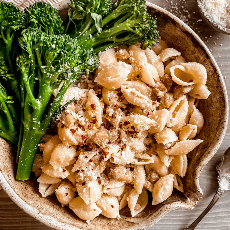 Pasta with sausage and broccolini is shown in a beige pottery bowl, a spoon is laying next to the bowl, a wooden bowl of extra parmesan cheese is in the top right corner and a red and white striped dish towel is in the top left corner.