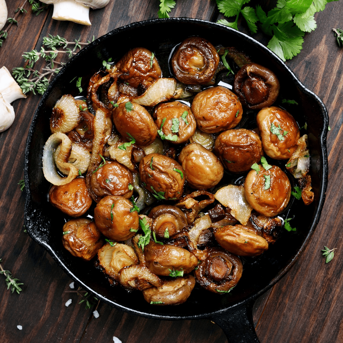 Cooked button mushrooms in a cast iron skillet on a wooden surface with whole white mushrooms and fresh parsley in the background.