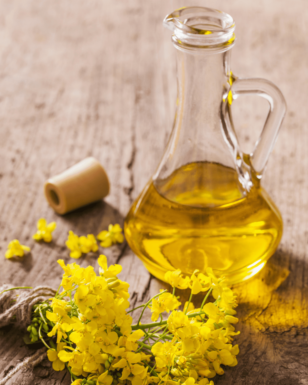 Glass bottle of canola oil with a cork and yellow flowers next to the bottle.