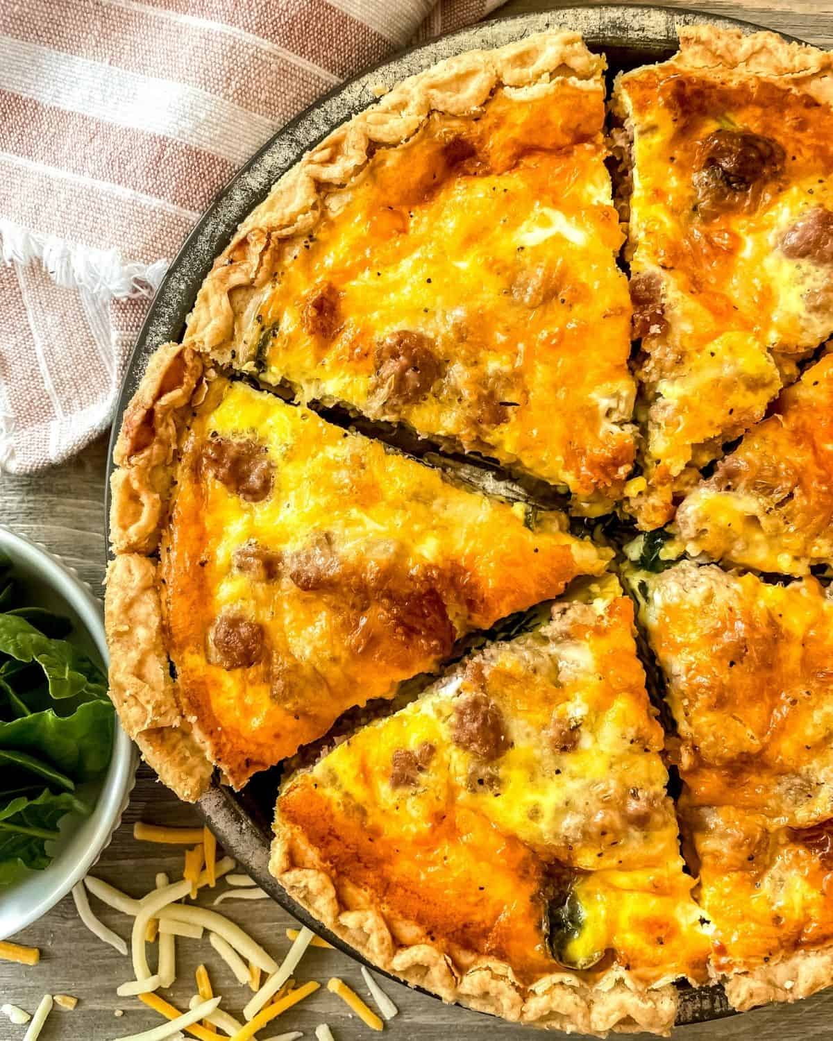 Sausage spinach quiche in a metal pie tin, a white bowl of spinach is on the side, sprinkling of cheddar cheese is below the quiche and a dish towel is in the top left corner.
