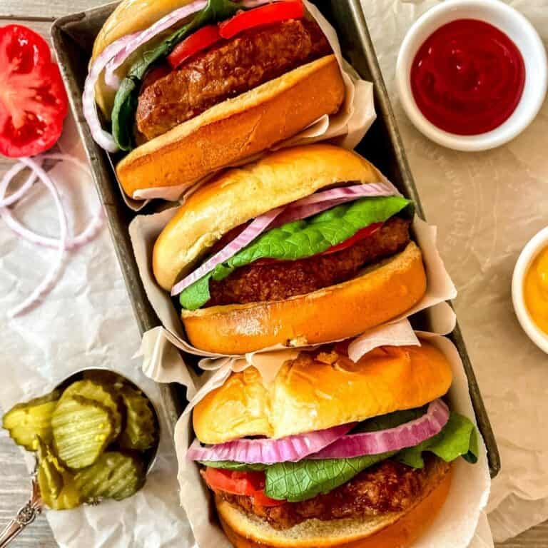 Air fryer frozen turkey burger in a bun with lettuce, onion and tomato, wrapped in parchment, stacked in a pile of three burgers, with ketchup, mustard and pickles on the side.