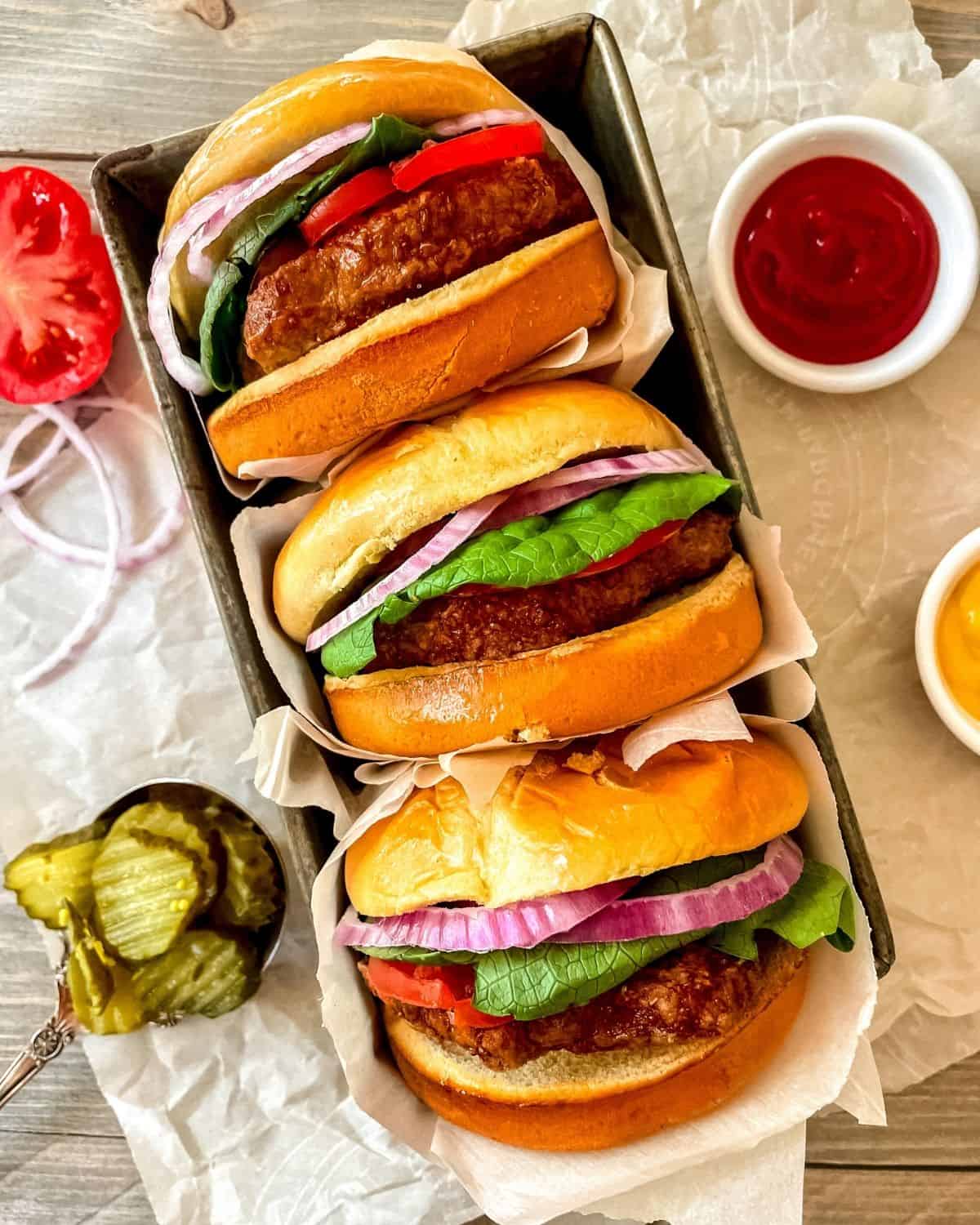 Air fryer frozen turkey burger in a bun with lettuce, onion and tomato, wrapped in parchment, stacked in a pile of three burgers, with ketchup, mustard and pickles on the side.