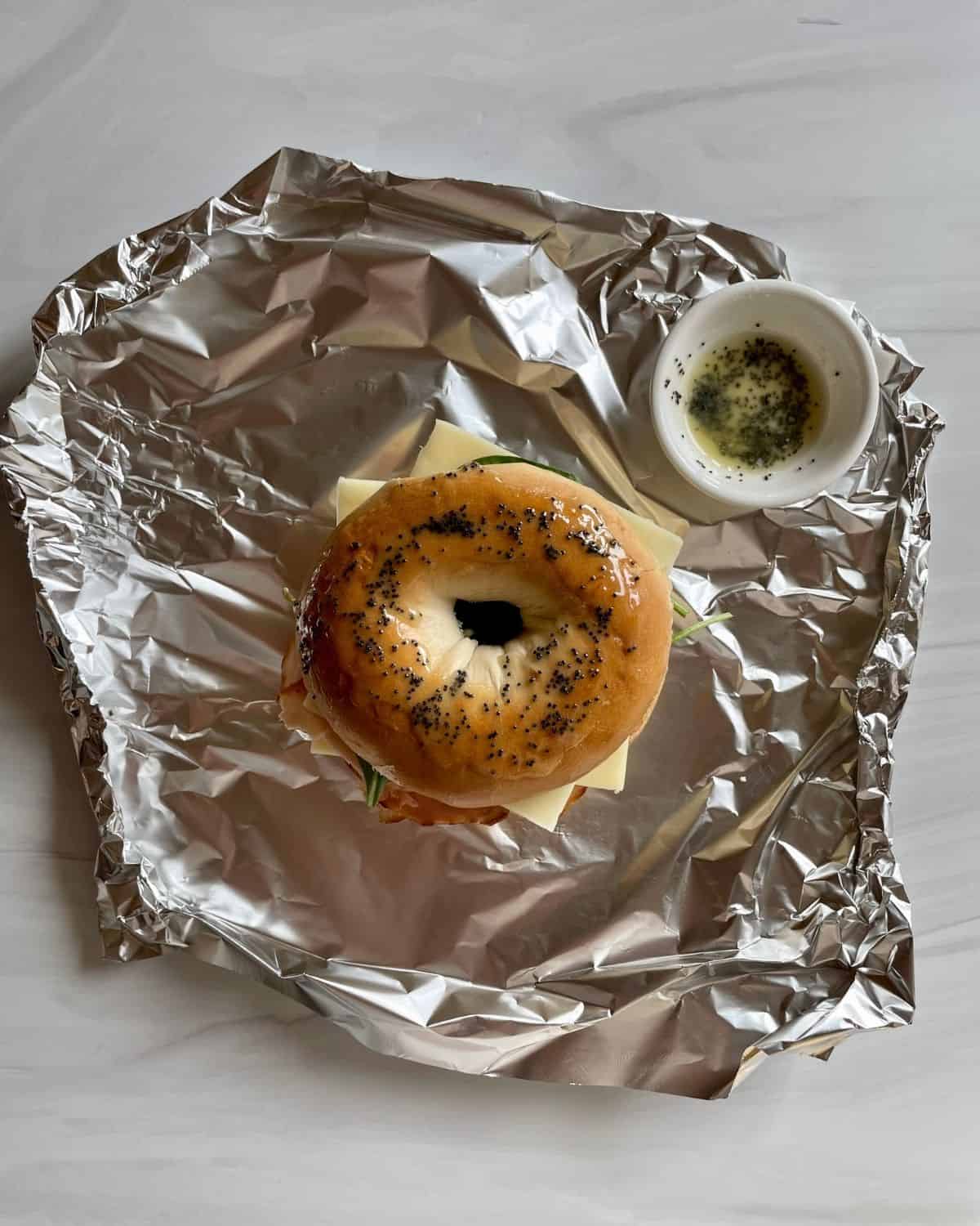 Bagel on tinfoil with poppy seed butter in a small bowl to the right.
