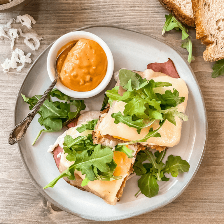 Open face turkey Rueben breakfast sandwich on a round grey plate, topped with arugula, TI dressing on the side and slices of bread and sauerkraut in the upper corners of the image.