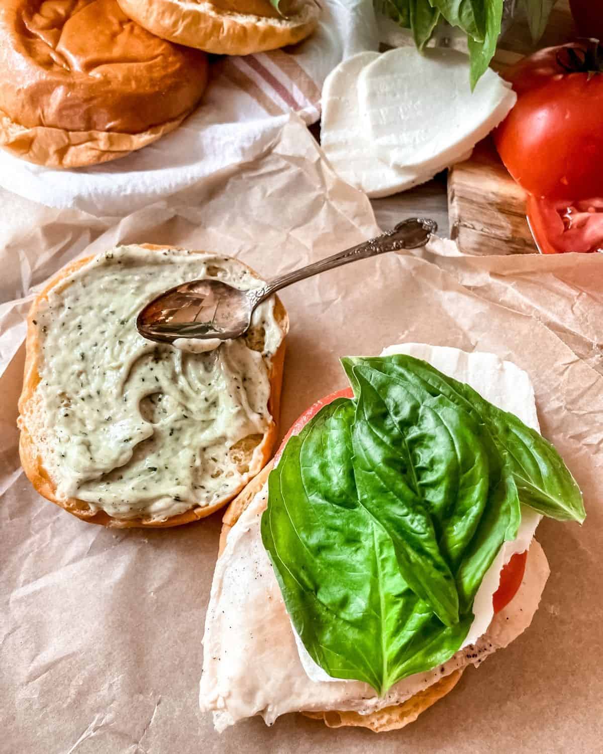 Open face chicken sandwich with pesto mayo spread on one side of the open bun, on parchment paper, extra buns, fresh mozzarella and tomatoes are seen in the background.