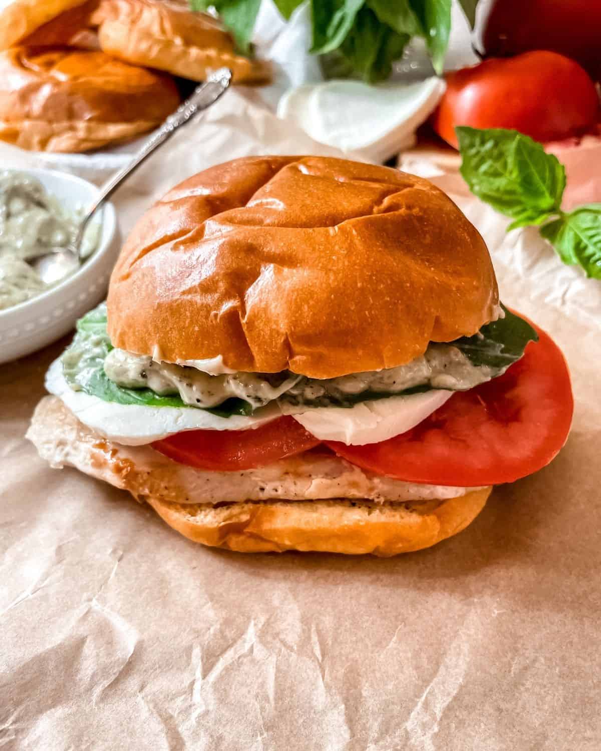 Caprese chicken sandwich on a brioche bun, on parchment paper, ingredients in the background.