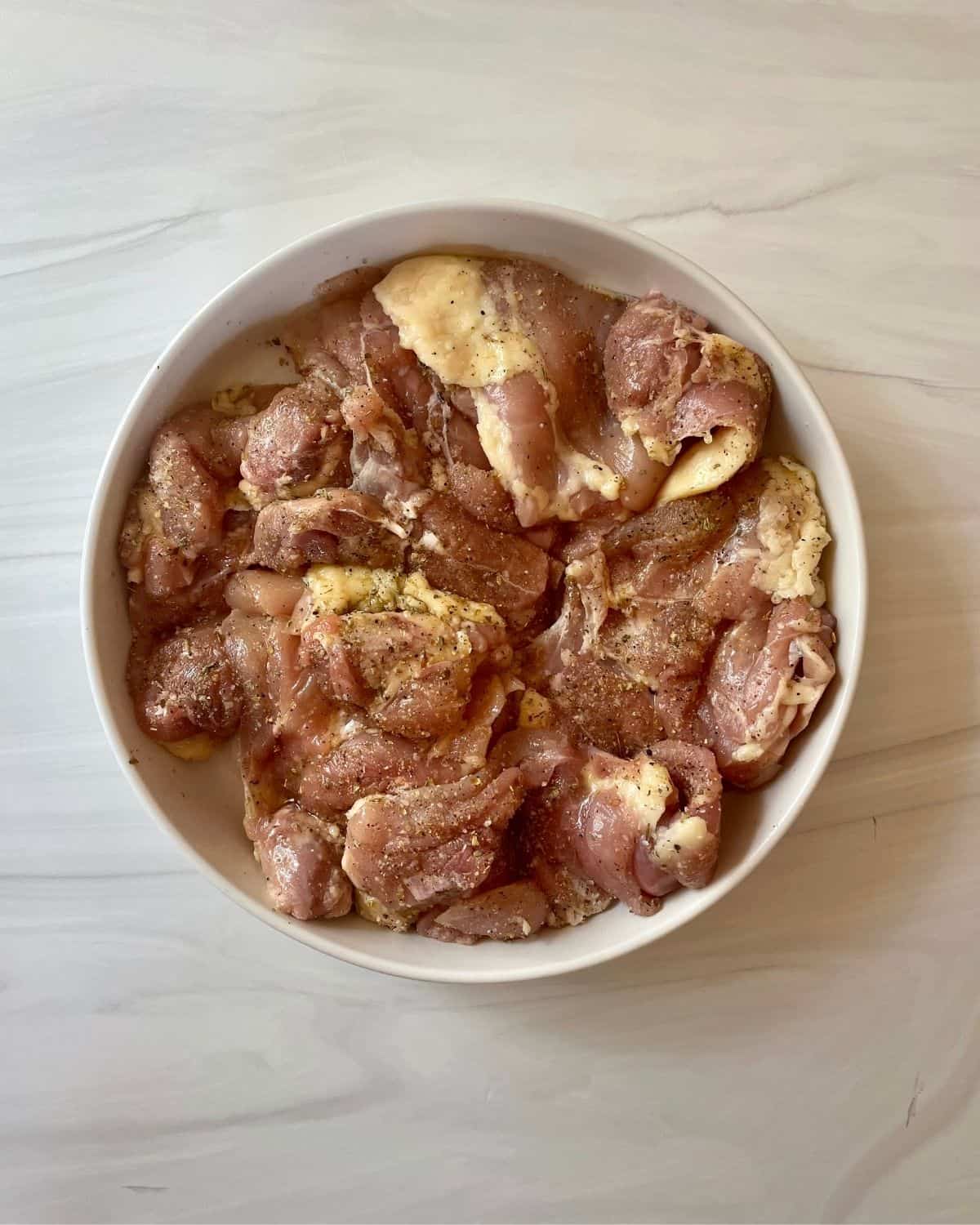 Raw boneless skinless chicken thighs with seasoning and olive oil in a round white bowl.
