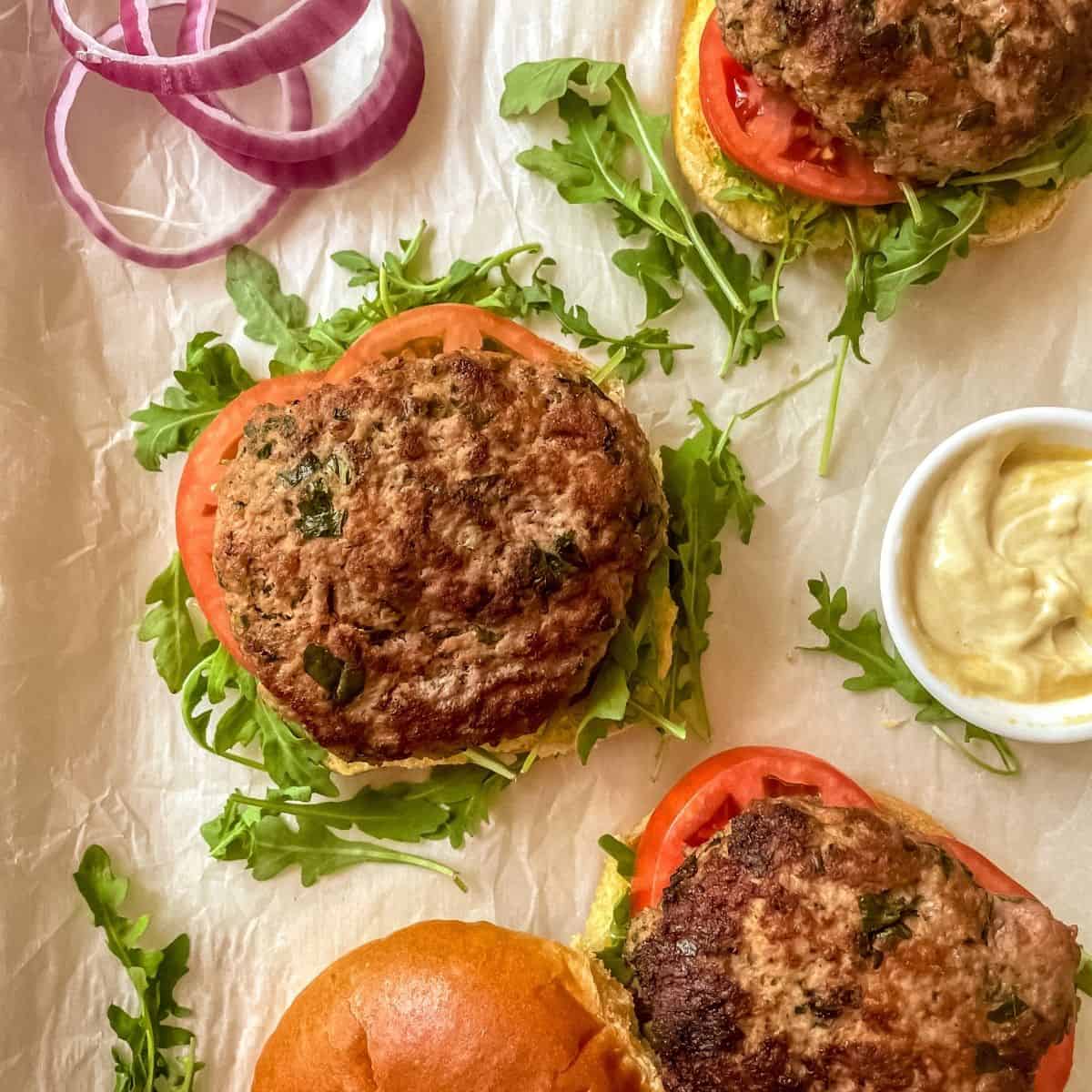 Three healthy turkey burgers on parchment paper, open face with arugula, red onion, sliced tomatoes, brioche buns and mustard.