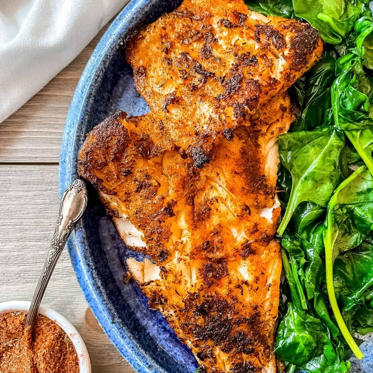 Blackened cod fillets on a round, blue plate, served with sautéed spinach. Blackening seasoning in the bottom left corner with a spoon. Top left corner has a white napkin.