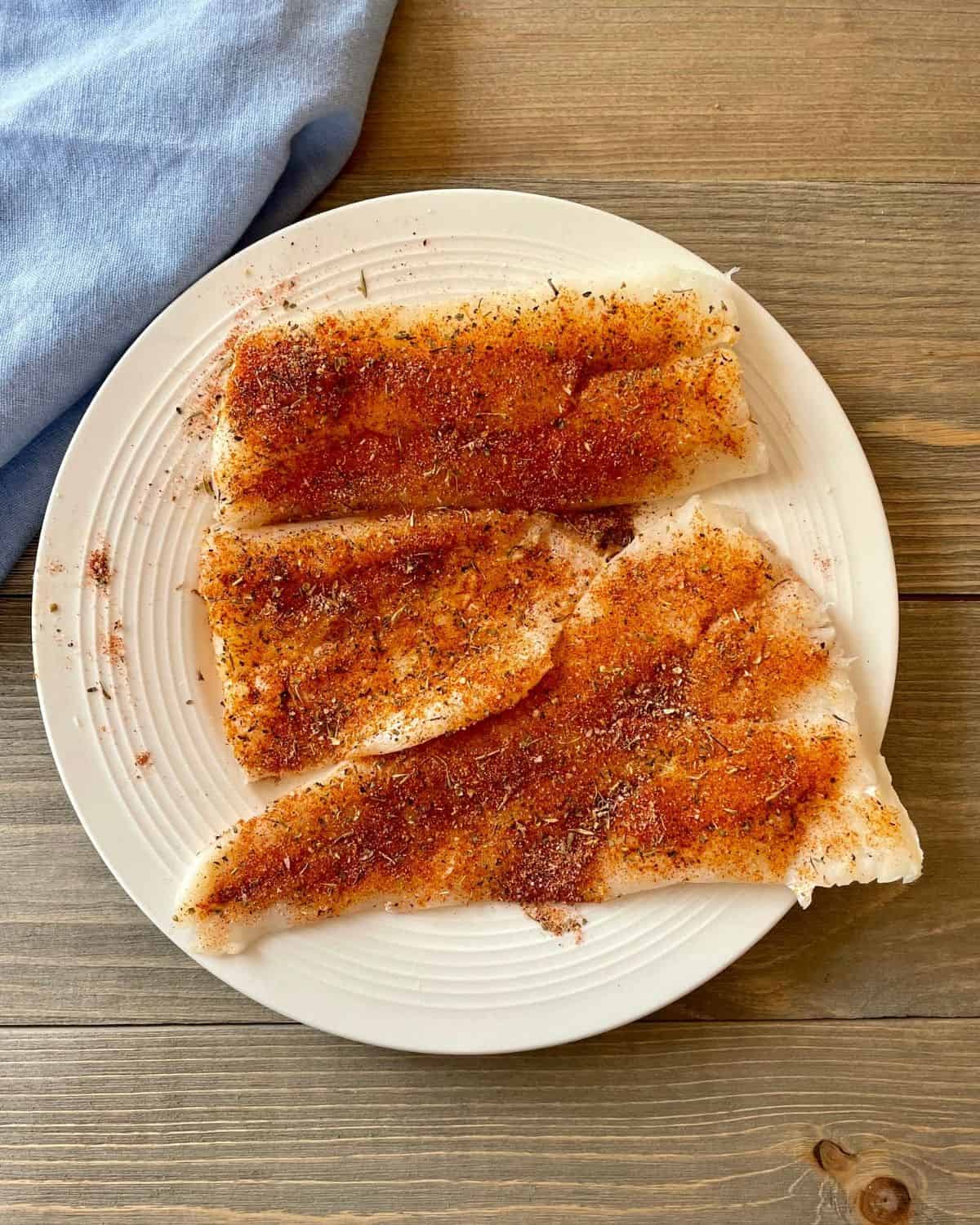 Raw cod fish fillets on a white plate, with blackening seasoning.
