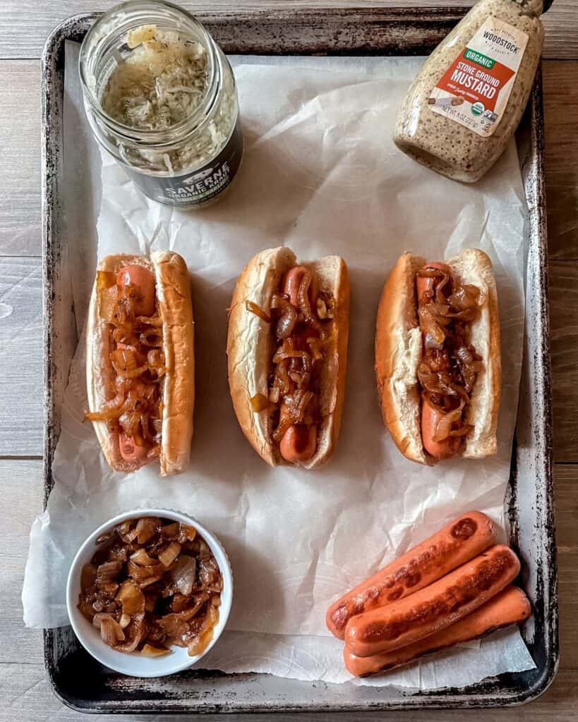 Three hot dogs topped with caramelized onions in buns, on a sheet pan, mustard in top right corner, sauerkraut in top left corner, caramelized onions in bottom left corner, cooked hotdogs in bottom right corner.