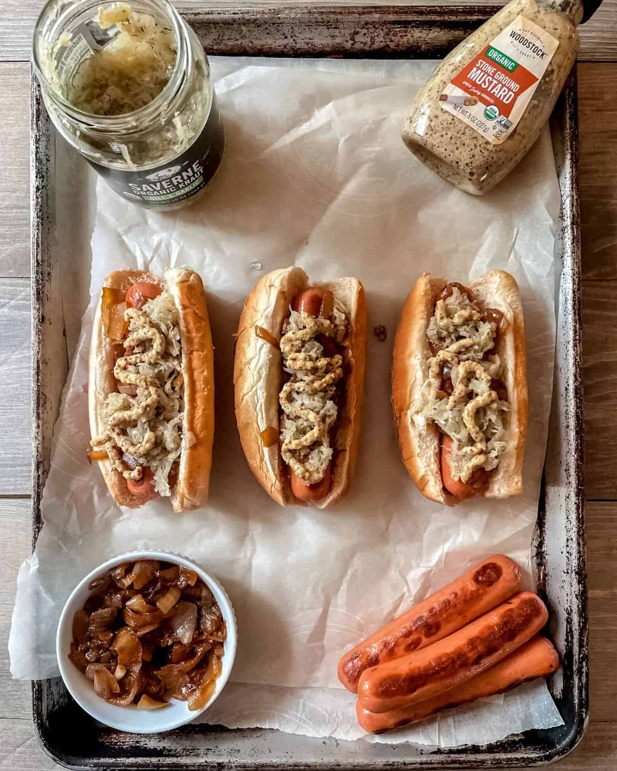 Three hot dogs topped with onions, sauerkraut and mustard in buns, on a sheet pan, mustard in top right corner, sauerkraut in top left corner, caramelized onions in bottom left corner, cooked hotdogs in bottom right corner.