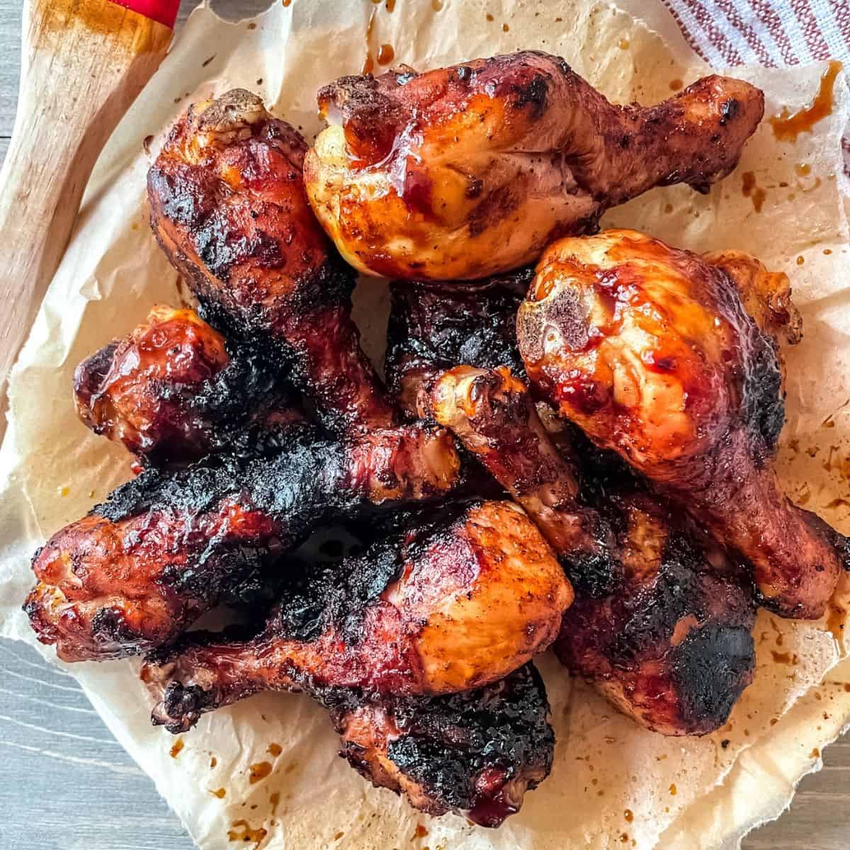 BBQ grilled chicken drumsticks recipe cooked and on a round plate, on top of brown parchment paper, a dish towel is in the corner and a basting brush with bbq sauce is in the top left corner.