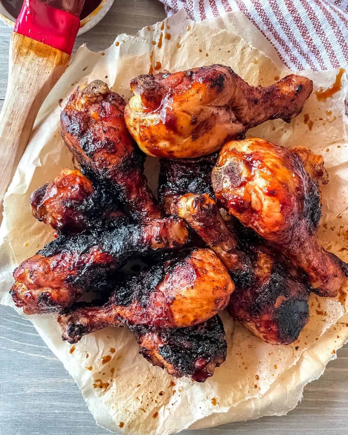 BBQ grilled chicken drumsticks recipe cooked and on a round plate, on top of brown parchment paper, a dish towel is in the corner and a basting brush with bbq sauce is in the top left corner.