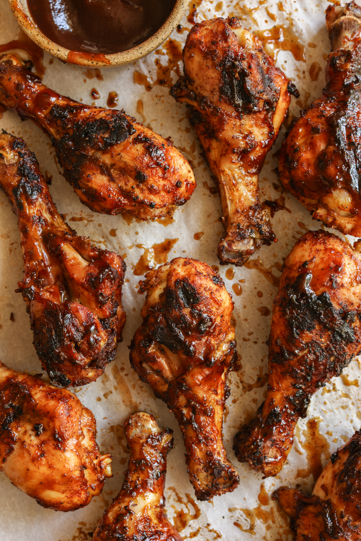 BBQ grilled chicken drumsticks smothered in BBQ sauce, on parchment paper, with a small dish of extra BBQ sauce on the side.