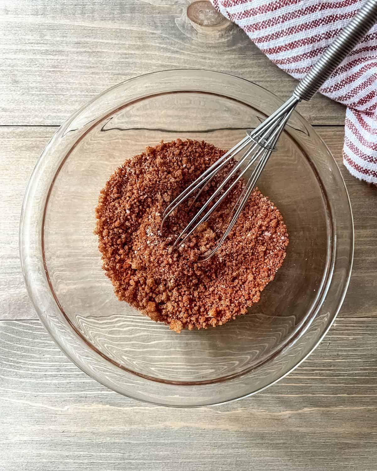 BBQ seasoning blend in a glass bowl with a whisk and a red and white striped dish towel in the corner.