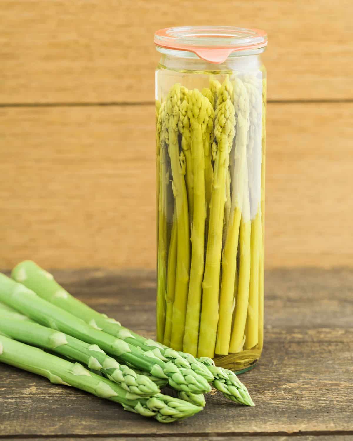 Canned asparagus in a glass jar, fresh asparagus laying next to the jar.