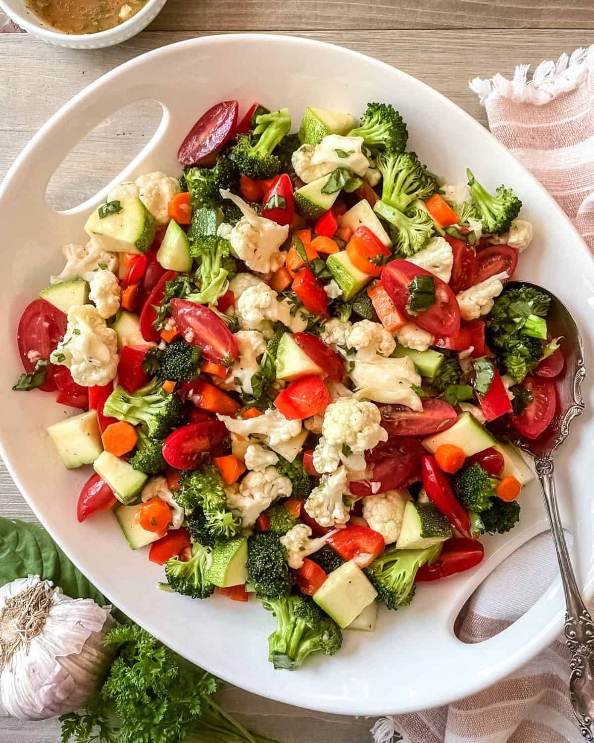 Marinated vegetable salad in a round white serving dish, with a large spoon, napkin on the side, bowl of marinade at the top, garlic and fresh herbs on the bottom left.