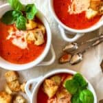 Roasted red pepper tomato soup in three white bowls with handles, on parchment paper, with croutons and fresh basil.
