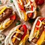 Four Italian sausages with cooked peppers and onions, in a bun on brown parchment paper, garnished with fresh parsley, a small bowl of mustard is in the bottom left corner and a spoonful of ketchup is in the top right corner.